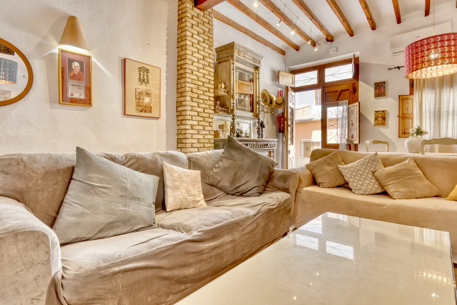 Salón con techos de vigas de madera, sofás beige con cojines, pared de ladrillo visto, lámpara colgante roja y puerta de entrada abierta que deja pasar luz natural.