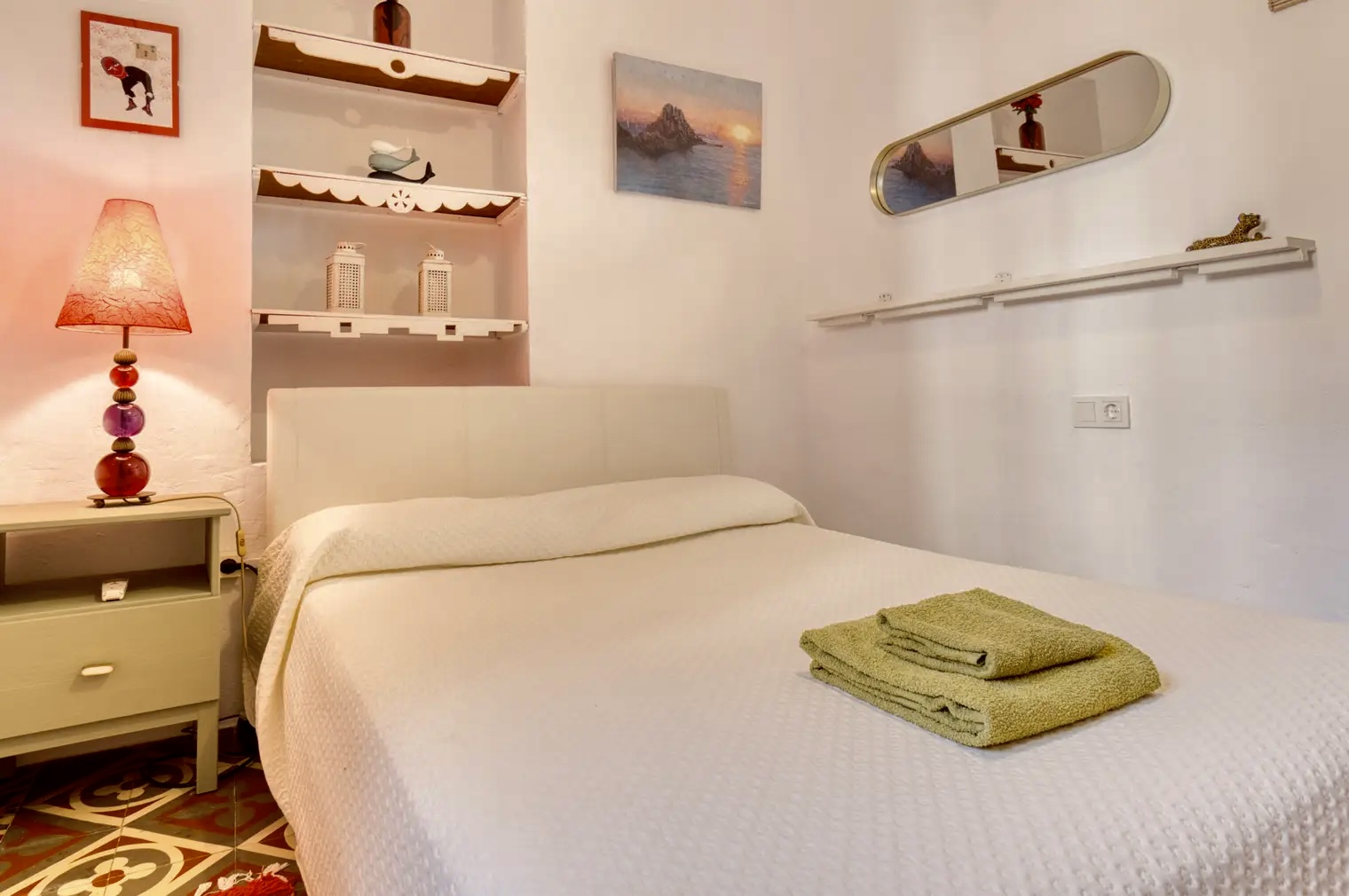 Dormitorio pequeño con cama doble vestida con colcha blanca, lámpara de mesa con pantalla roja y estanterías decorativas sobre la cabecera.