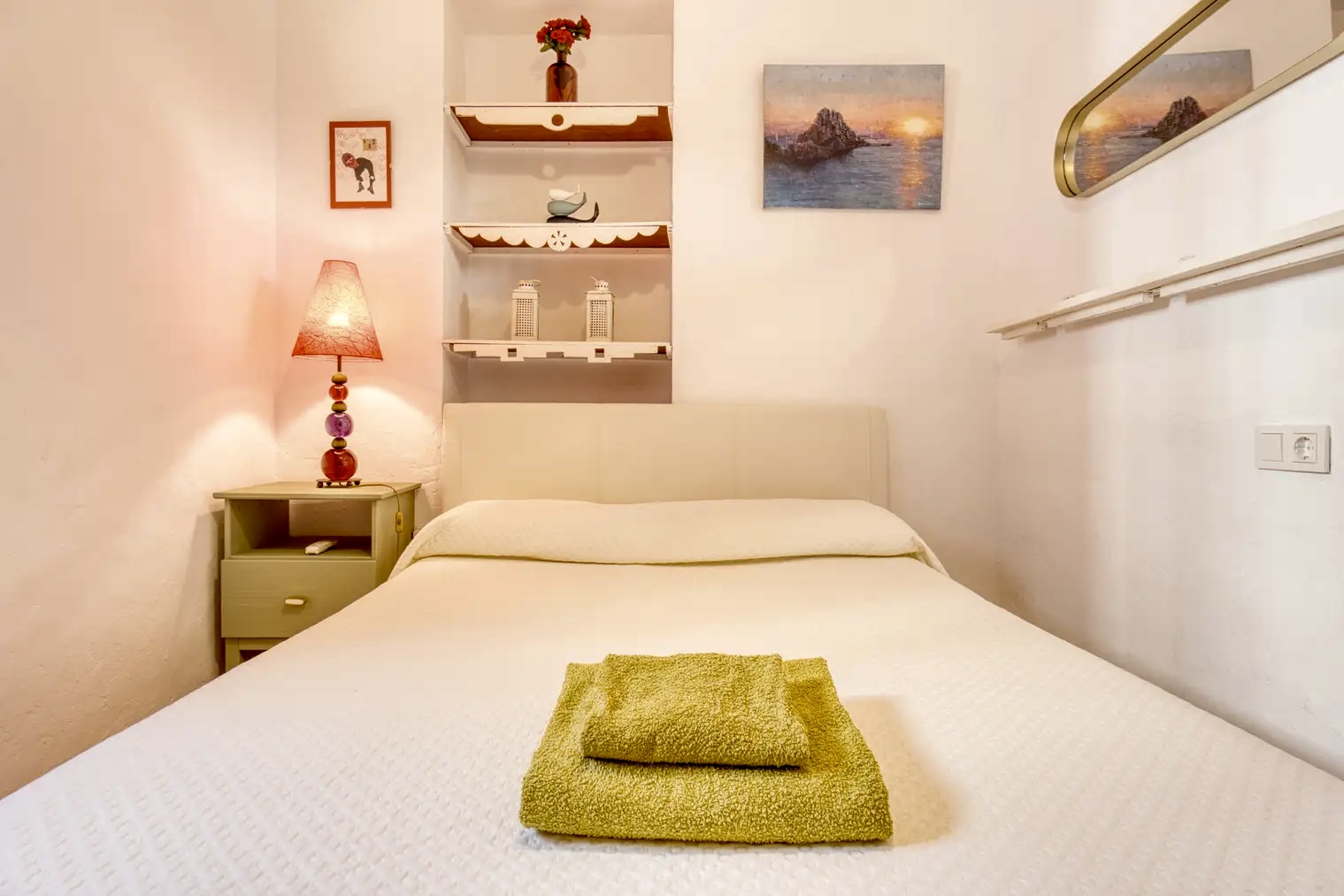 Dormitorio pequeño con cama doble vestida en blanco, lámpara de mesa con pantalla roja y estanterías decorativas sobre la cabecera.