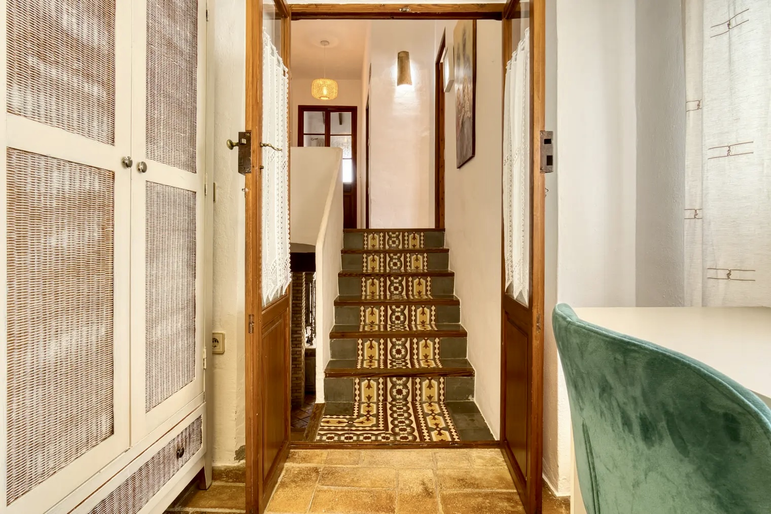 Pasillo interior con escaleras decoradas con mosaicos, puertas de madera con cortinas, armario con rejilla y silla verde junto a una mesa blanca bajo una ventana con cortinas claras.