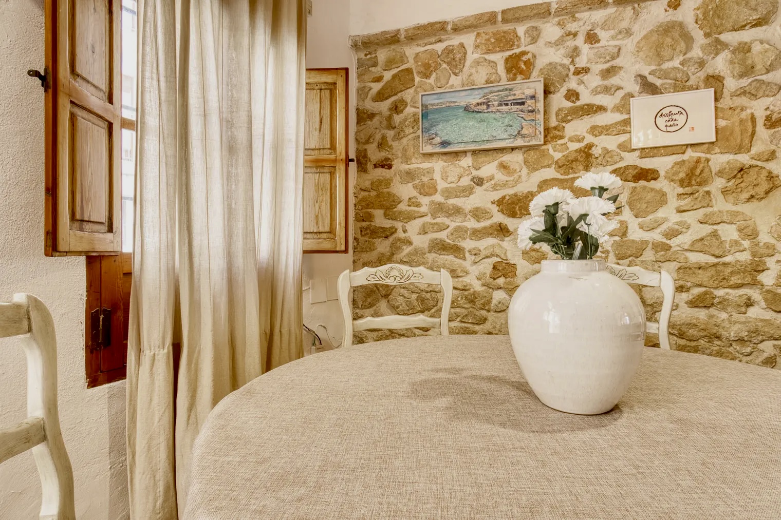 Comedor rústico con mesa redonda cubierta con mantel beige, jarrón blanco con flores, sillas de madera tallada y pared de piedra vista decorada con cuadros.