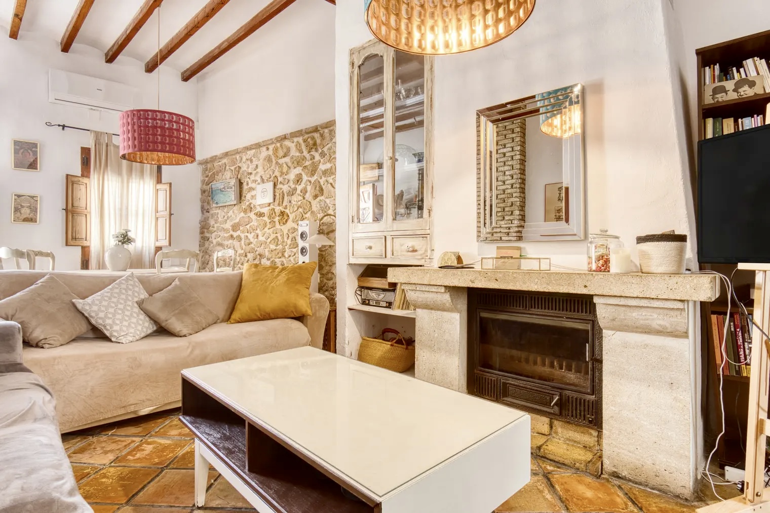 Salón rústico con sofás beige, chimenea de piedra, mesa de centro blanca, pared de piedra vista, estanterías con libros y lámparas colgantes decorativas.
