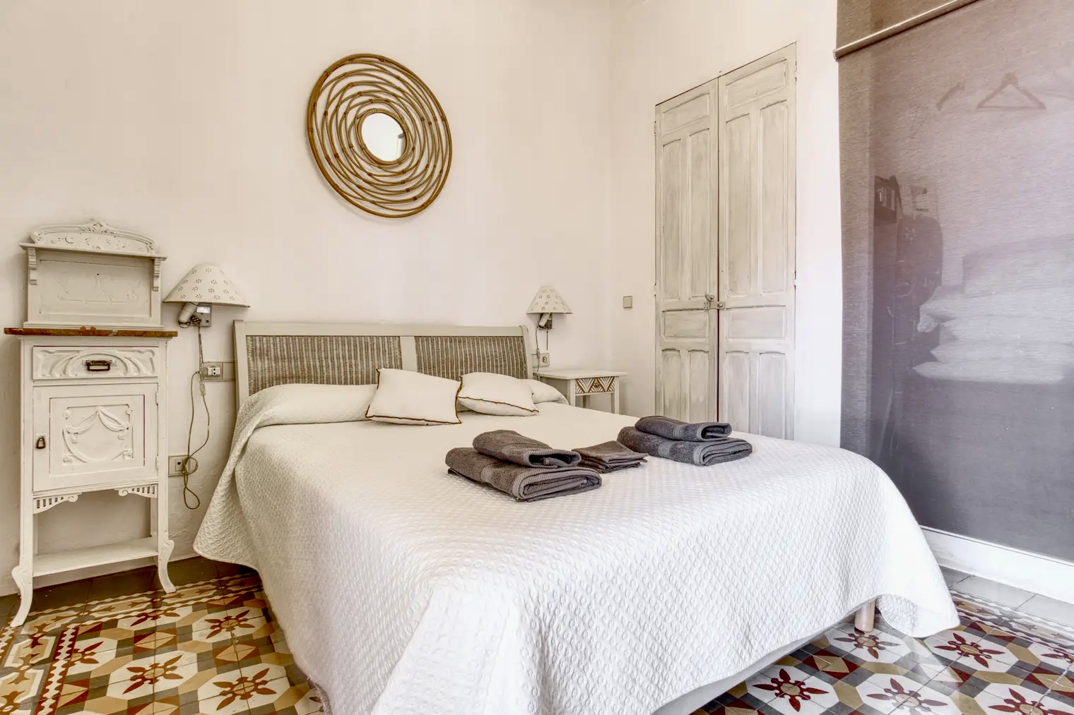 Dormitorio con cama doble vestida con colcha blanca, muebles vintage blancos, armario rústico y espejo redondo decorativo en la pared.