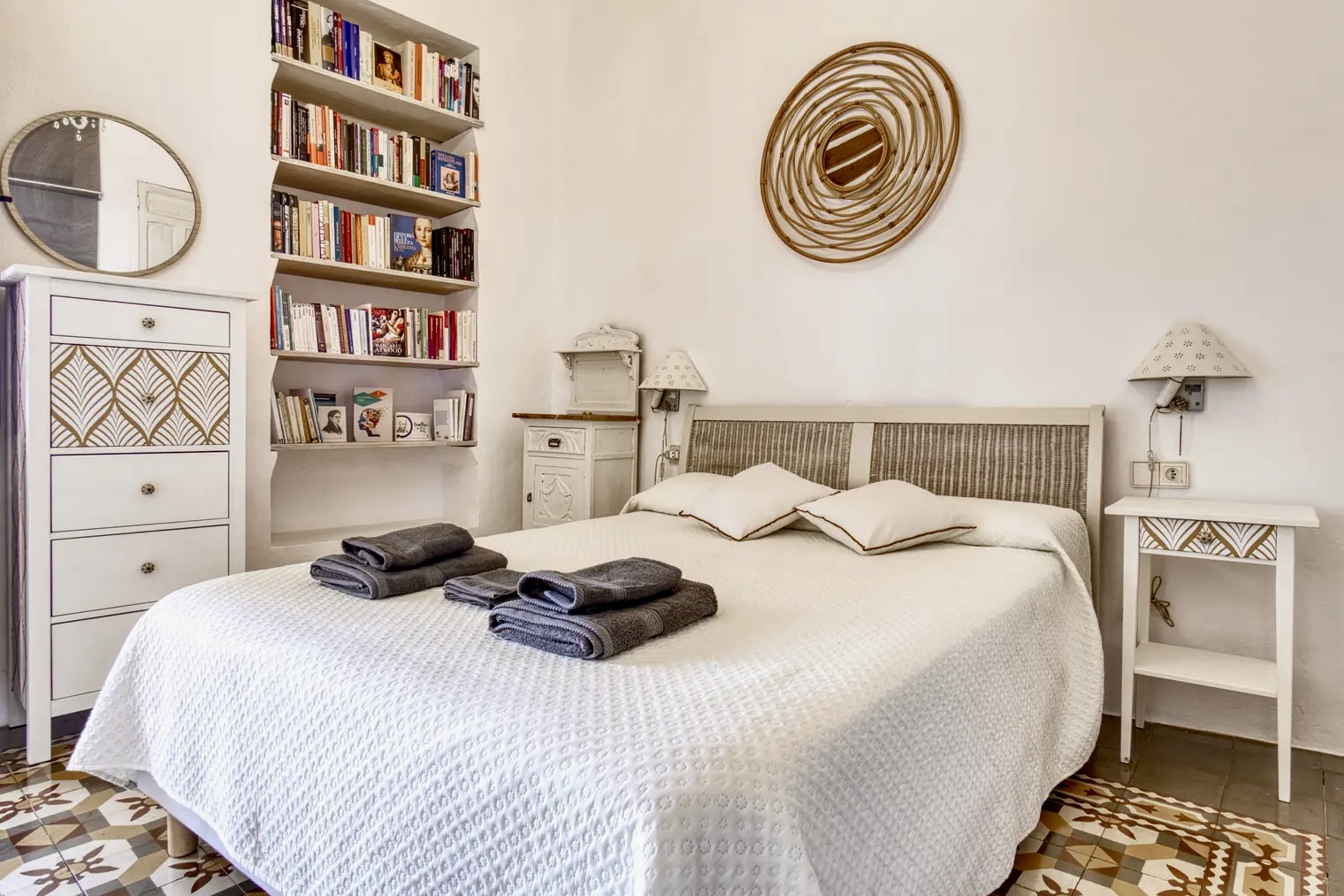 Dormitorio decorado con cama doble, estantería llena de libros, cómodas blancas con detalles dorados y adorno circular de mimbre en la pared.