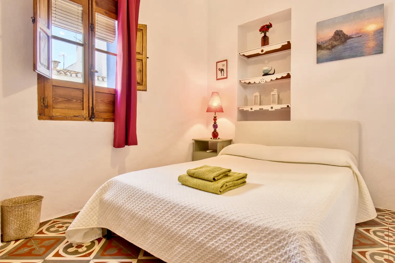 Dormitorio acogedor con cama doble, colcha blanca, toallas verdes dobladas, lámpara de mesa, estantería decorativa y ventana de madera con cortinas rojas.