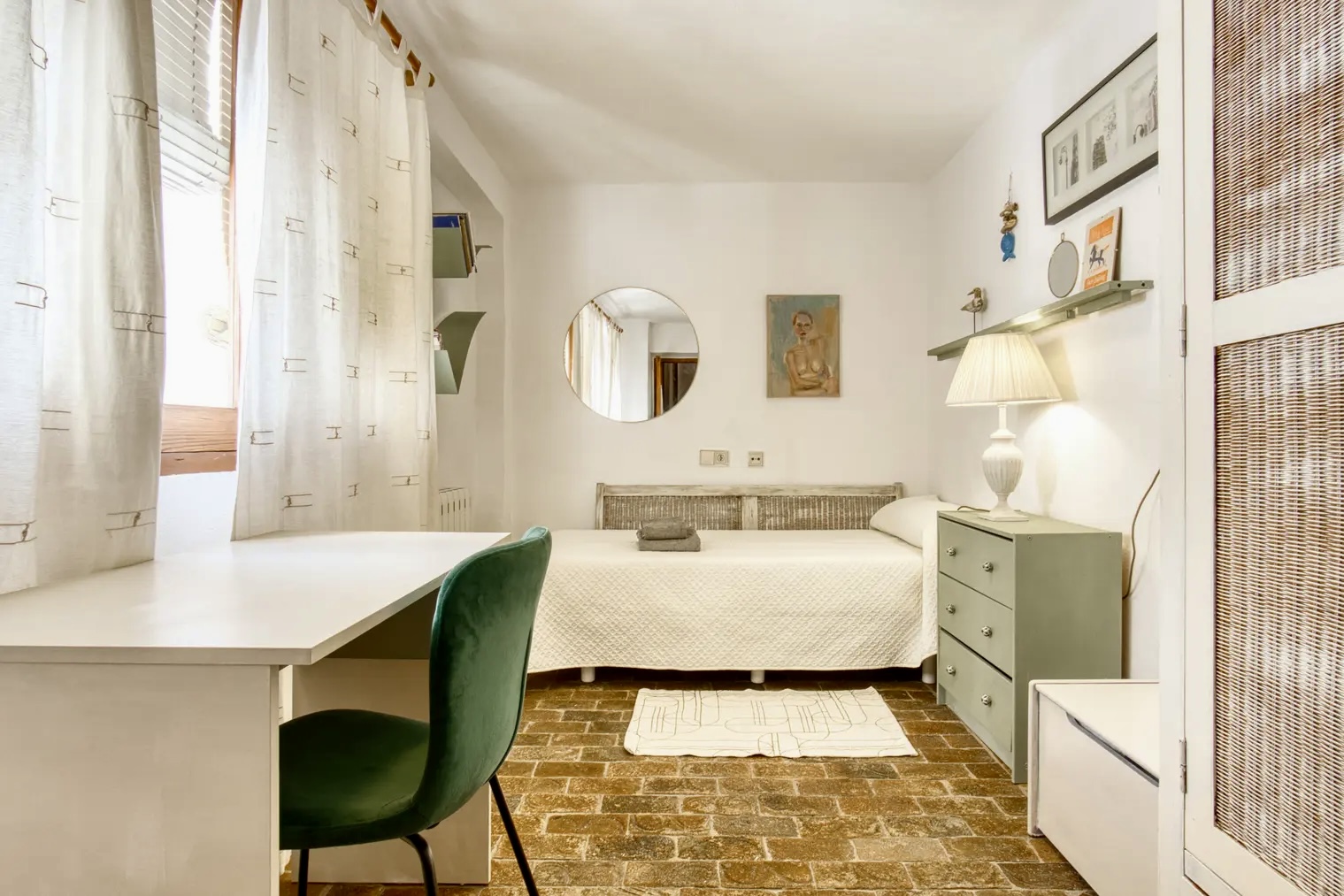 Dormitorio luminoso con cama individual, escritorio blanco con silla verde, cómoda verde con lámpara, espejo redondo en la pared y suelo de ladrillo rústico.