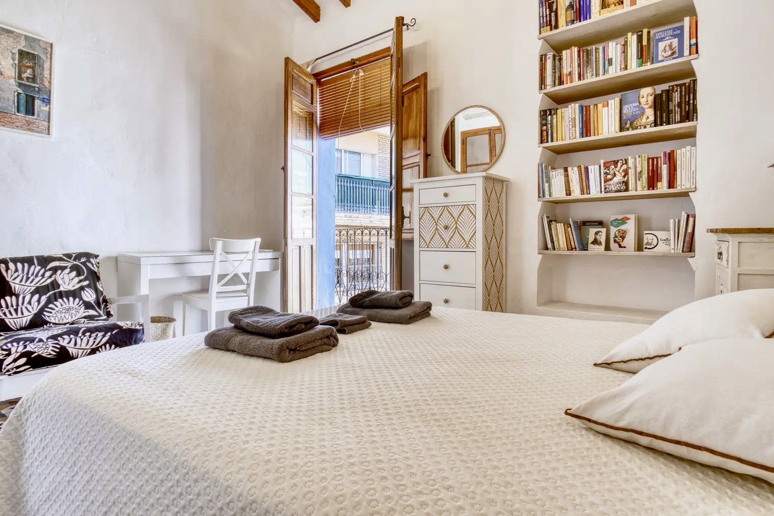 Dormitorio luminoso con cama doble, toallas dobladas, escritorio blanco, estantería con libros, cómoda decorativa y puertas de madera que dan a un balcón exterior.