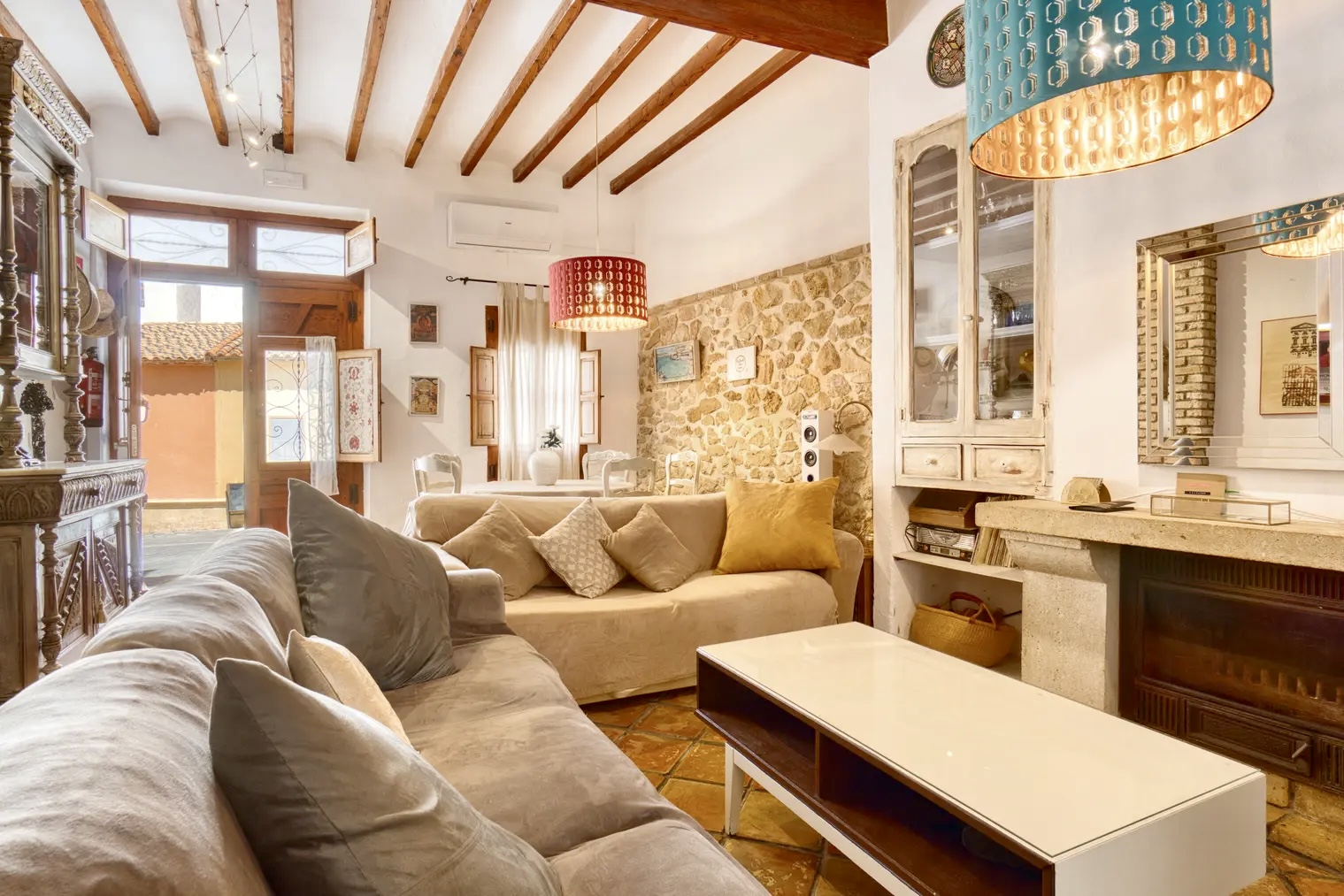 Acogedor salón rústico con sofás beige, lámparas colgantes, chimenea, mesa blanca de centro y pared de piedra, iluminado por luz natural que entra por la puerta abierta.