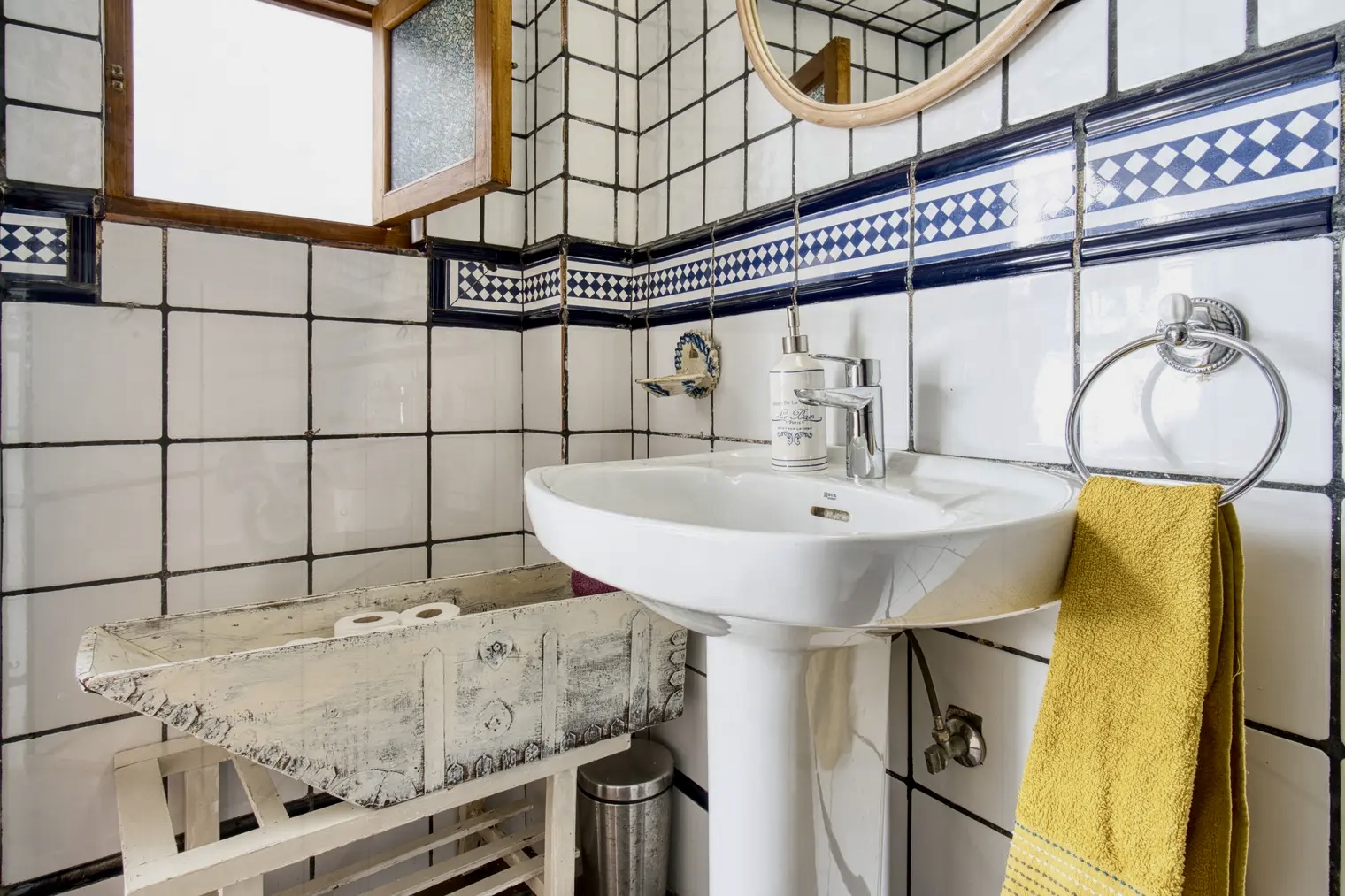 Baño casa rural Les Avies con azulejos blancos y detalles en azul, lavabo con dispensador de jabón, toalla amarilla, espejo redondo y estantería rústica.