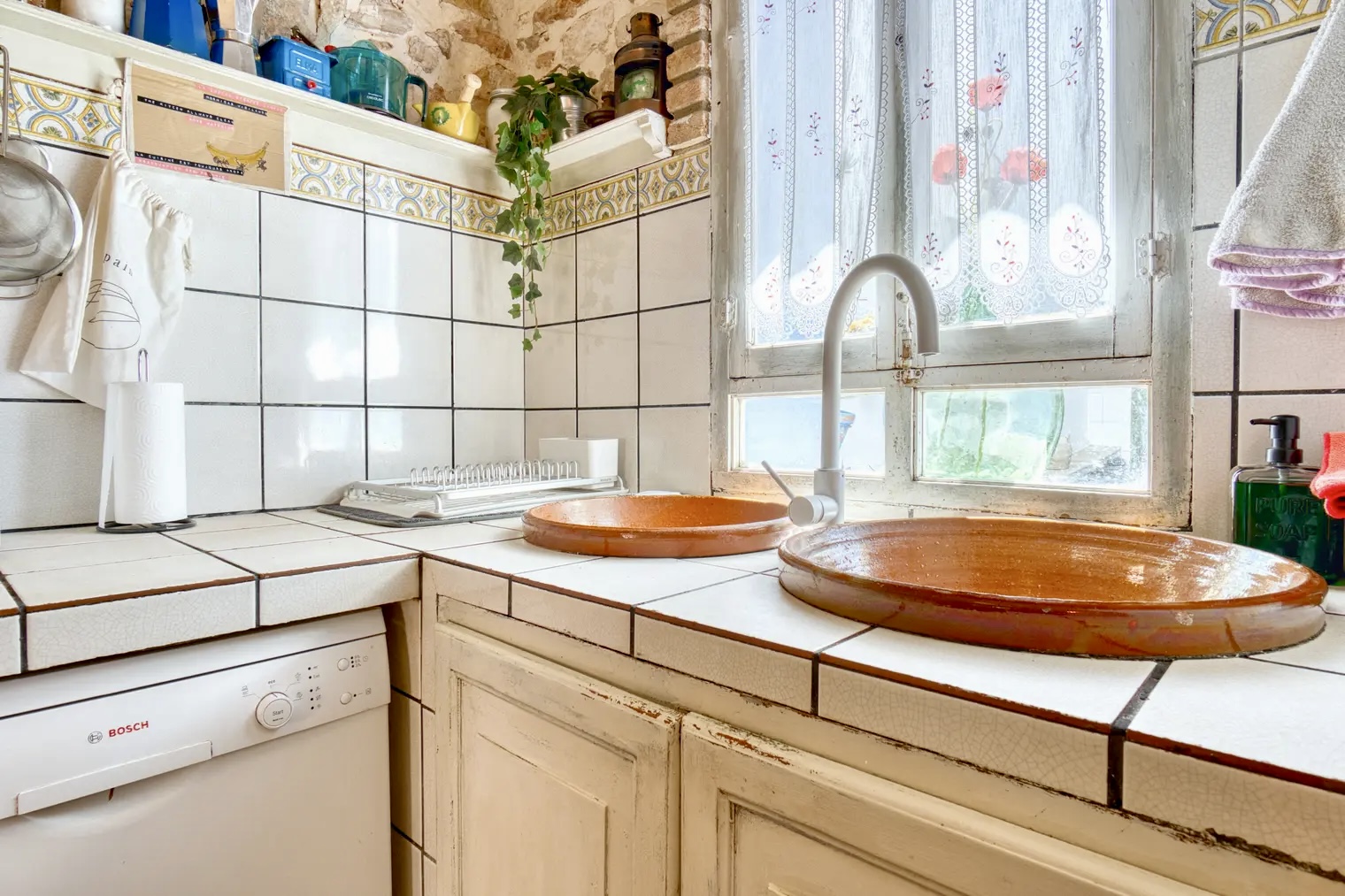 Cocina rústica con fregadero doble de barro, lavavajillas Bosch, encimera de azulejos blancos y ventana con cortinas bordadas que deja pasar la luz natural.