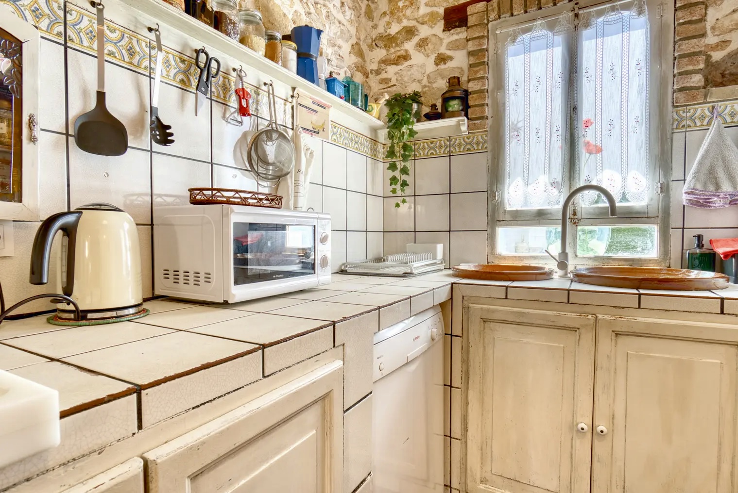 Acogedora cocina rústica con azulejos blancos, utensilios colgados, microondas, hervidor eléctrico, fregadero doble de barro y ventana con cortinas bordadas.