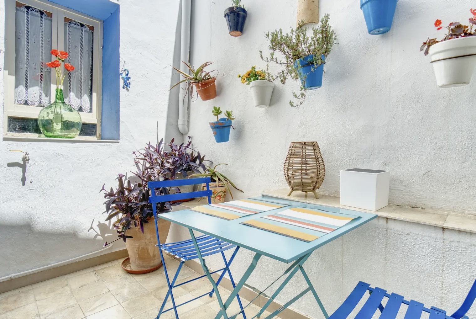 Acogedor patio blanco con mesa y sillas azules, decorado con plantas en macetas de colores colgadas en la pared y detalles rústicos junto a una ventana con cortinas bordadas